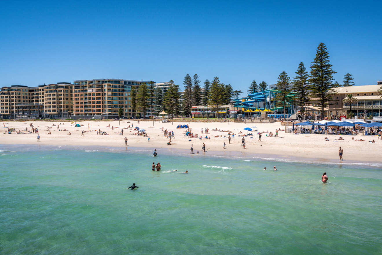 People enjoy a sunny day at a beach with gentle waves, surrounded by buildings and trees in the background.