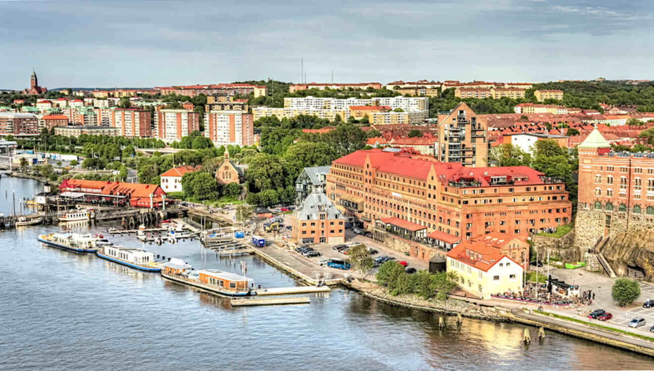 Aerial view of a coastal city with red-roofed buildings, a waterfront with docks, and boats moored along the shore. Green spaces and urban areas are visible in the background.