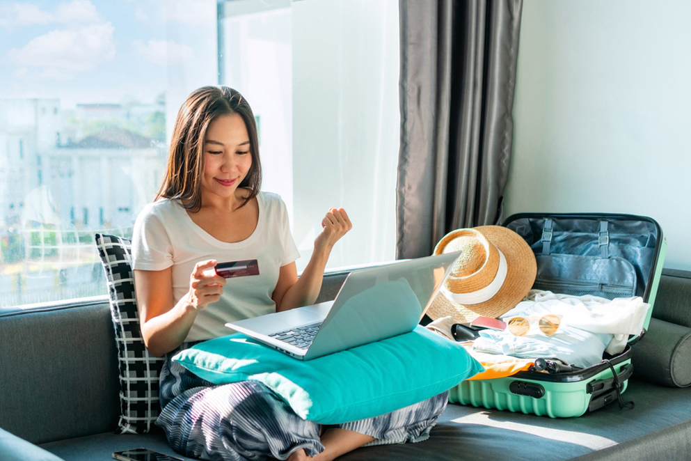 A woman sitting on a sofa uses a laptop, holding a credit card. A packed suitcase with clothes and a straw hat is beside her. Natural light enters through a window.