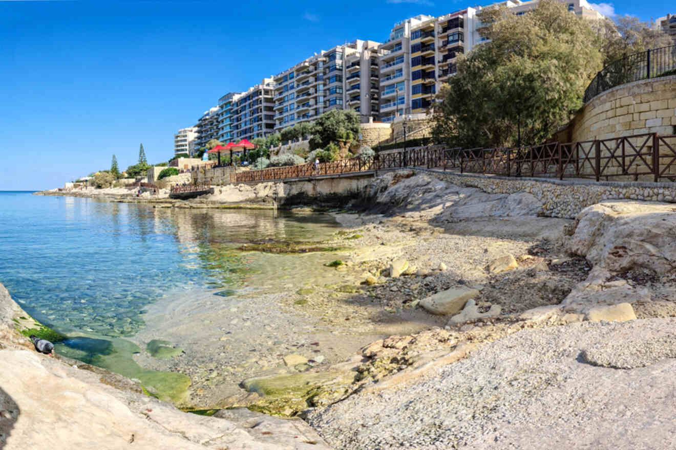 Coastal view of rocky shoreline with clear blue water and a distant line of multi-story buildings under a clear sky.