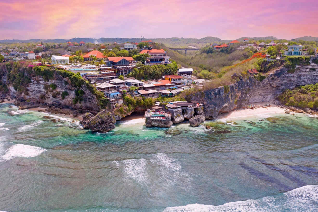 Aerial view of a coastal cliff with buildings, surrounded by ocean waves and greenery under a pinkish sky.