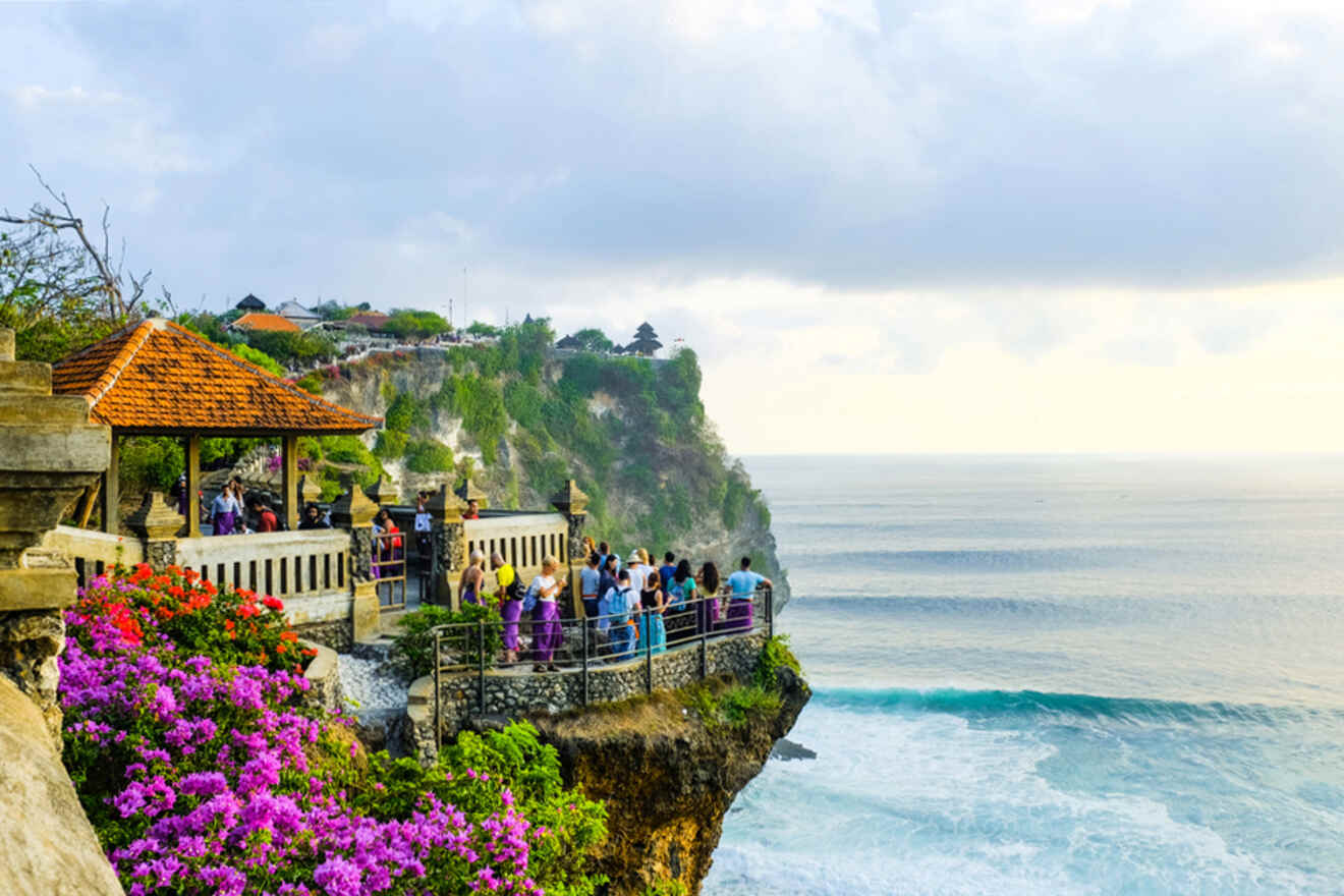 A group of people gathered at a cliffside viewpoint overlooking the ocean, with vibrant purple flowers in the foreground and a distant coastline under a cloudy sky.