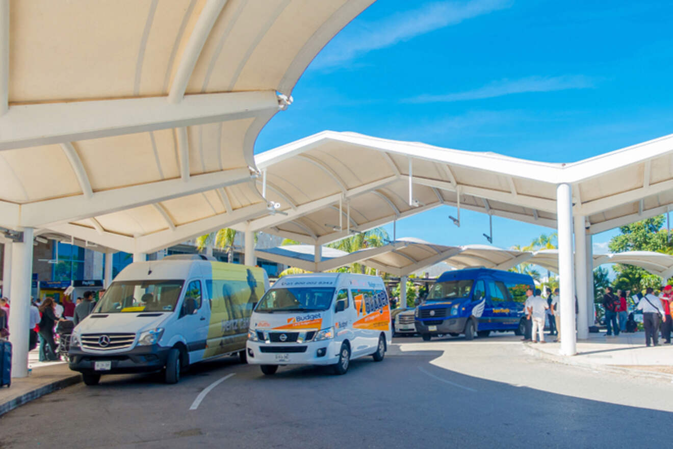 Several colorful shuttle vans parked under a white canopy at an outdoor transportation hub on a sunny day.