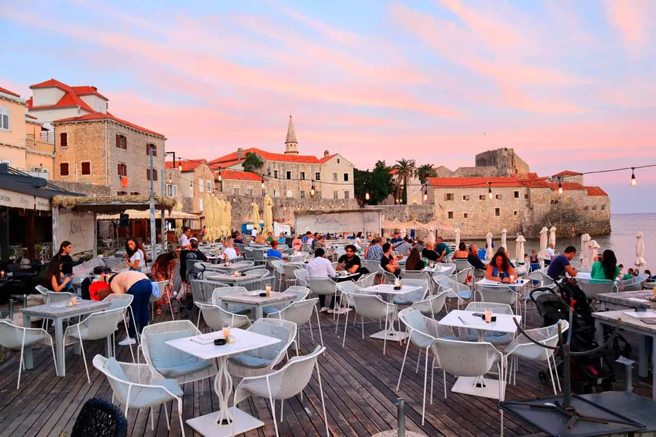 People sit at outdoor tables on a wooden deck, surrounded by historic stone buildings, with a pink sunset sky in the background.