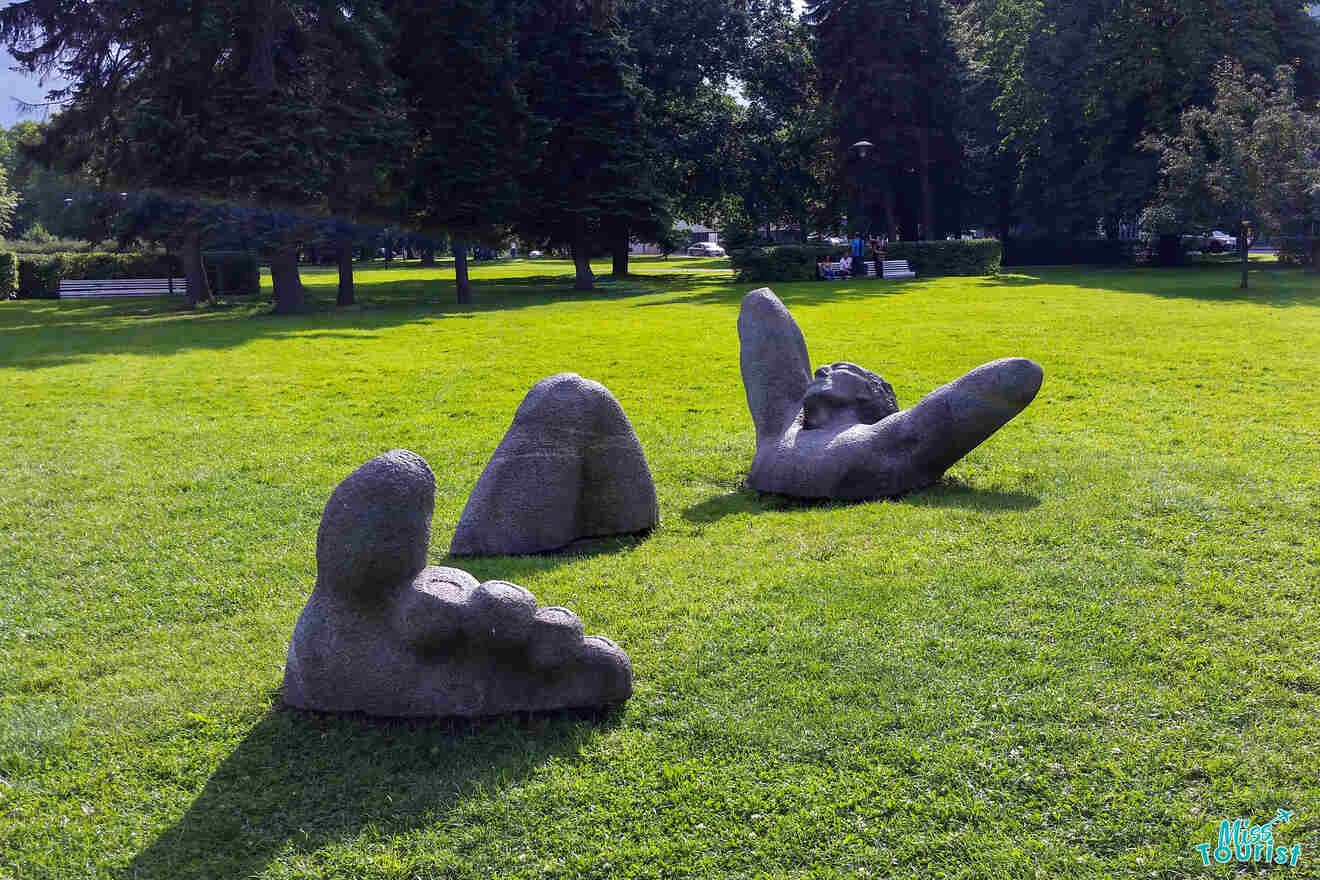 Stone sculpture of a partial figure emerging from the grass, depicting a head, arms, and feet, set in a park with trees in the background.