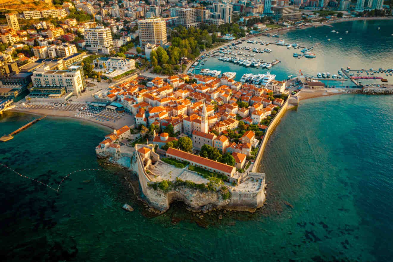 Aerial view of a coastal town with orange-roofed buildings, surrounded by a stone wall. Nearby, a marina is visible with boats docked, and modern buildings are in the background.