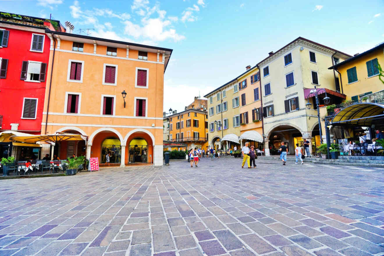 A colorful town square with vibrant buildings and a cobblestone plaza. People are walking and there are outdoor seating areas with umbrellas. The sky is partly cloudy.