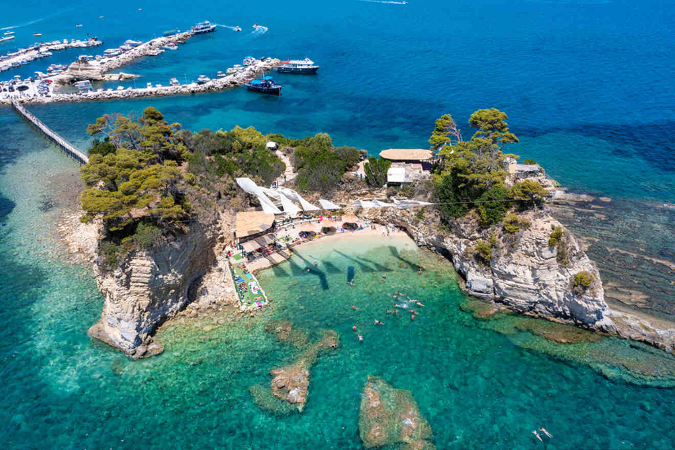 Aerial view of a small island with a beach, sunbathers, and umbrellas. Surrounded by clear blue water and boats docked nearby. Rocky formations and greenery visible.