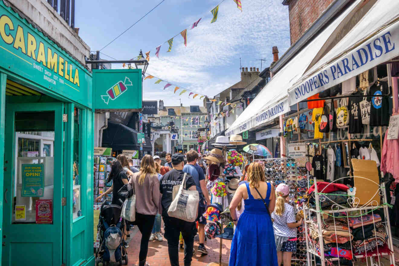 Busy outdoor market street with people shopping. Brightly colored shopfronts and stalls display various items for sale. Bunting decorates the area, and the sky is clear and blue.