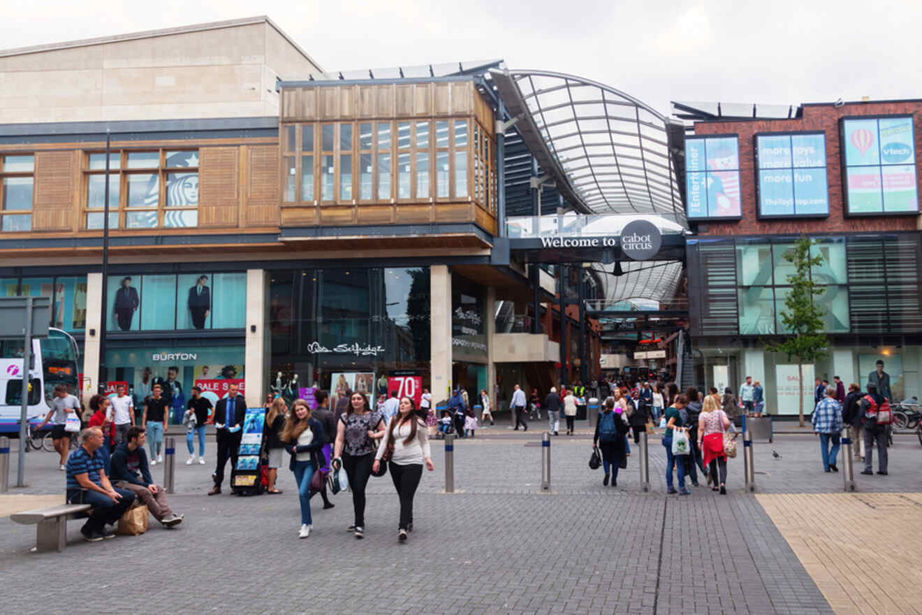 People walking and sitting in a busy shopping area with modern buildings and a "Welcome to Cabot Circus" sign overhead.