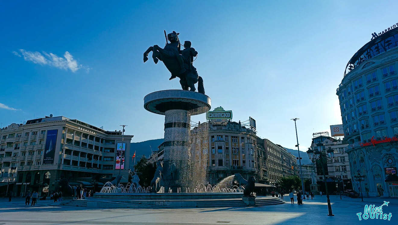 A large equestrian statue atop a fountain in a city square, surrounded by historic buildings under a clear blue sky.