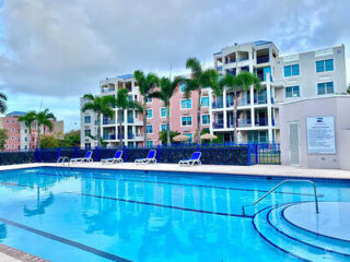 Outdoor swimming pool with blue lounge chairs, surrounded by palm trees and pink and white apartment buildings under a cloudy sky.