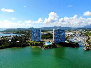 Aerial view of two tall apartment buildings near a marina, surrounded by green water and lush landscape under a partly cloudy sky.