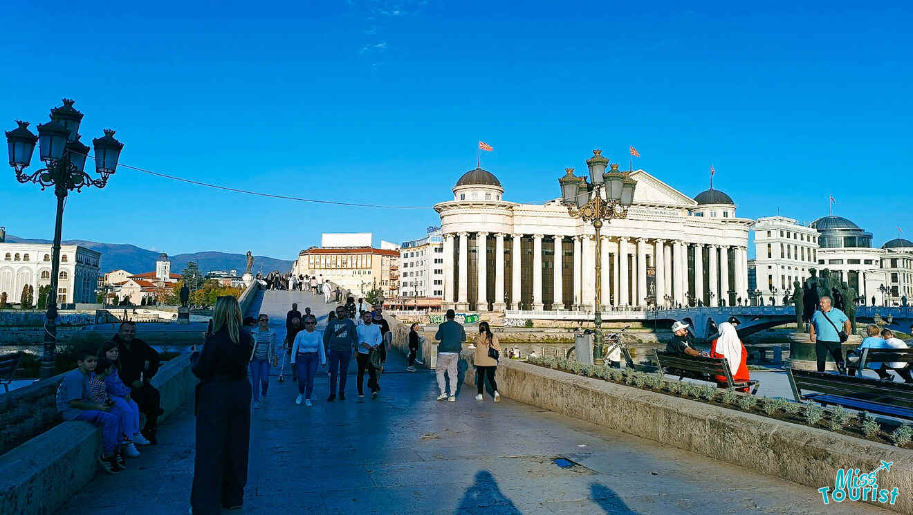 People walking on a bridge with a neoclassical building and street lamps in the background.