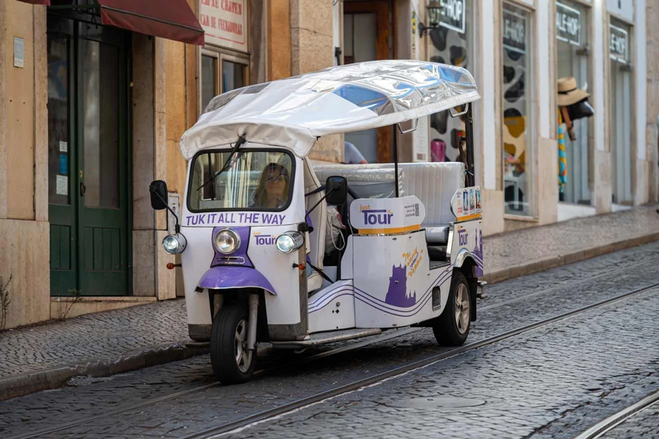 A white and purple tuk-tuk with "Tuk It All The Way" signage is parked on a cobblestone street near shopfronts.