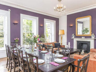 Dining room with a long table set for eight, surrounded by dark wooden chairs, a purple accent wall, large windows, a fireplace with a mirror above, and a chandelier overhead.