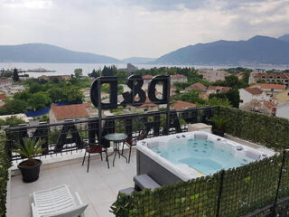 Rooftop terrace with a hot tub, table, and chairs, overlooking a town and distant mountains with a body of water. A metal railing has large D&B letters. Greenery surrounds the area.