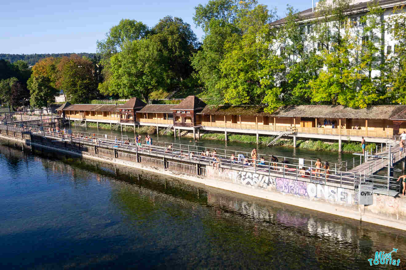 Riverside area with a wooden walkway and huts, surrounded by trees. People are strolling along the path on a sunny day.