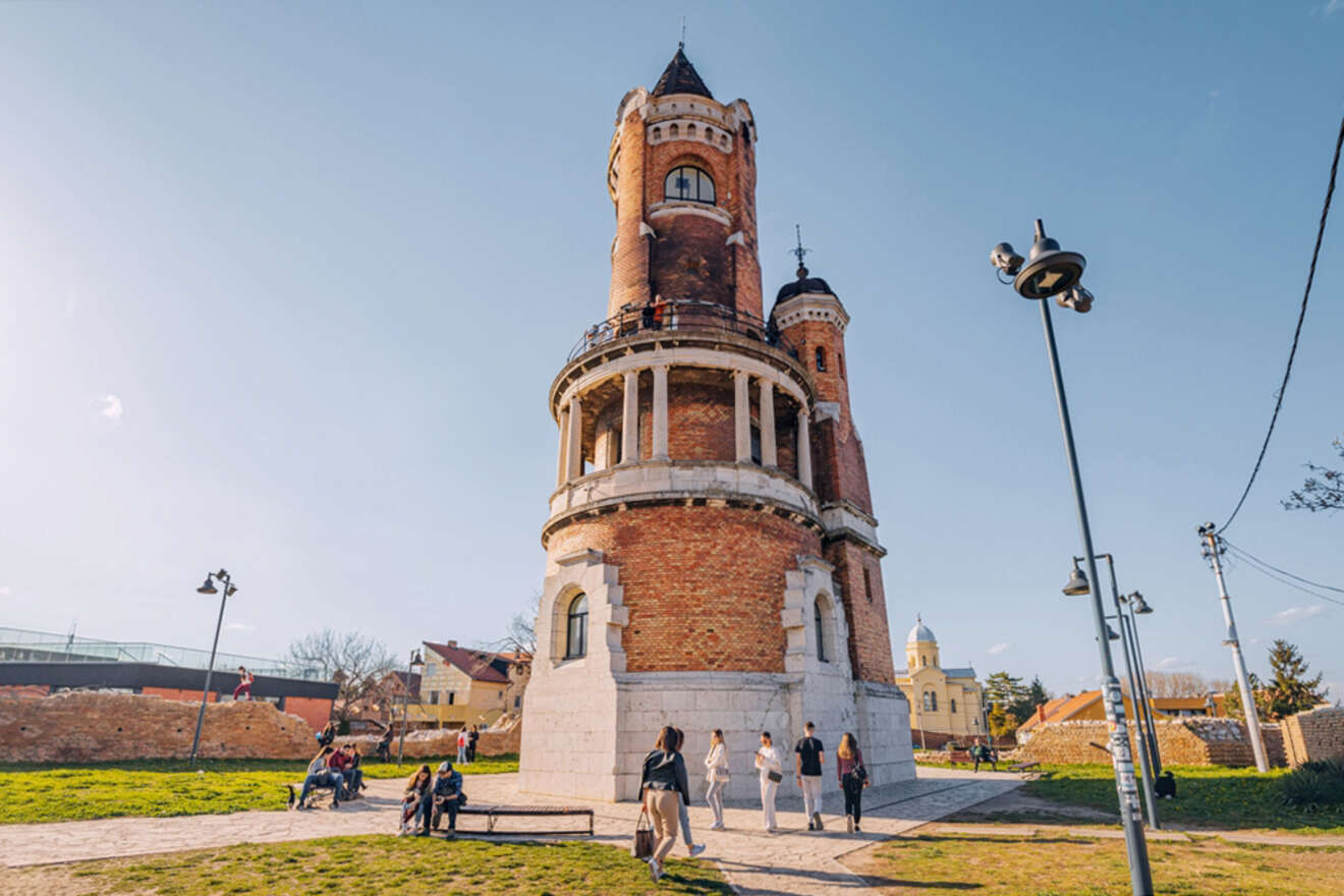 A tall, cylindrical brick tower with turrets stands in a park, with people walking and sitting around it on a sunny day.