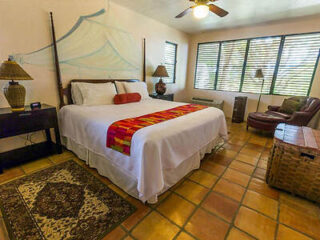 A cozy bedroom with a large bed, colorful throw, two side tables with lamps, an armchair, a wicker basket, and terracotta tiled floor, lit by natural light from the window.