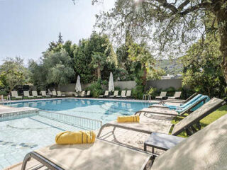 Outdoor pool area with sun loungers, umbrellas, and surrounding trees. A yellow towel is on one lounger.