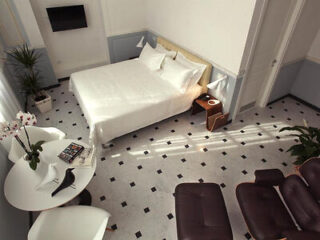 A modern, minimalist bedroom with a double bed, wall-mounted TV, round table with chairs, a lounge chair, and potted plants on a tiled floor with black accents.