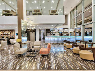 Modern hotel lobby with stylish seating, patterned flooring, and a bar area. Large windows and bright lighting create an open atmosphere.