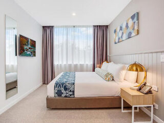 A modern bedroom with a neatly made bed, decorative pillows, a bedside table with a lamp and phone, artwork on the wall, a mirror, and a window with sheer curtains.