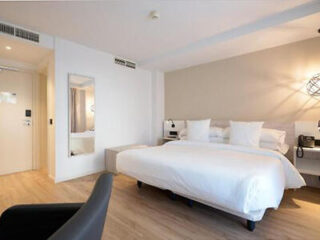 Modern hotel room with a king-size bed, white bedding, wall-mounted bedside lights, a large mirror, and a black chair on a light wooden floor.