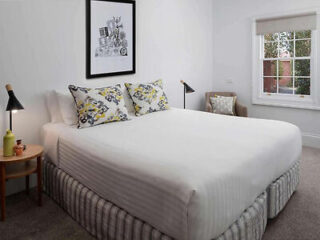 A tidy bedroom with a large bed, two decorative pillows, two bedside lamps, a framed picture above the bed, and a window with a view. There's also a small side table and a chair.