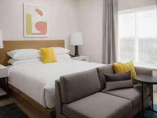 A neatly made bed with white linens and yellow pillows is beside a grey sofa. There is a side table with a lamp and abstract art on the wall. Natural light enters through the window.