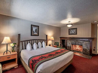 Hotel room with a large bed, multiple pillows, and a red bed runner. A lit fireplace is on the right. There are lamps on both sides of the bed and a framed picture on the wall.