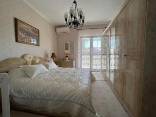 A cozy bedroom with a large bed, chandelier, wardrobe with mirrored doors, and a window draped with sheer curtains.