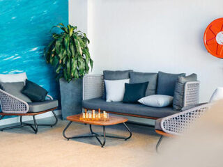 A modern seating area with a gray sofa and chairs, a wooden coffee table with a chess set, a large plant, blue wall art, and a red wall-mounted fire hose.