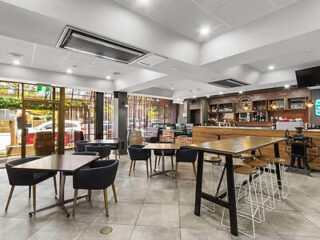 A modern cafe interior with wooden tables and black chairs. A long counter with bar stools is on the right, and large windows provide natural light.