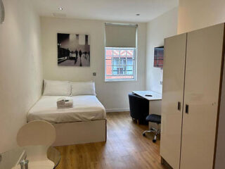 A minimalist bedroom with a bed, wardrobe, desk, and chair. Wall art and a window with blinds are visible. The floor is wooden.