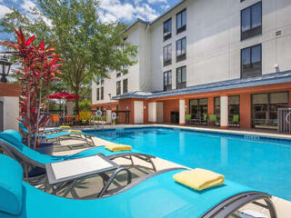 Outdoor pool area with blue lounge chairs and umbrellas, next to a building with multiple windows and surrounded by trees.