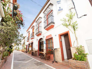 A narrow street lined with white buildings featuring balconies and outdoor seating. Plants and flowers adorn the facades. A sign reads "Hostal Restaurante El Gallo.