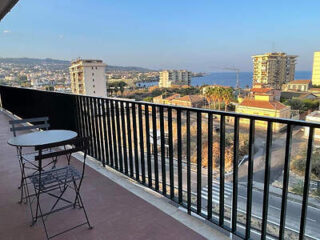 Balcony with a round table and chairs overlooks a cityscape with buildings and a view of the sea in the distance.