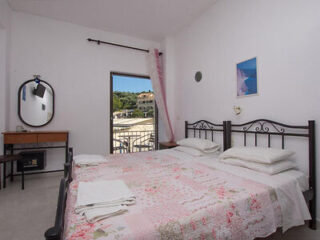 A minimal bedroom with a metal-framed double bed, a floral quilt, bedside table, mirror, and open window revealing a view of buildings and greenery outside.