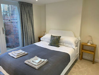 A tidy bedroom features a neatly made bed with a gray comforter, a plant-themed pillow, folded towels, two wooden side tables, lamps, and a window with closed curtains.