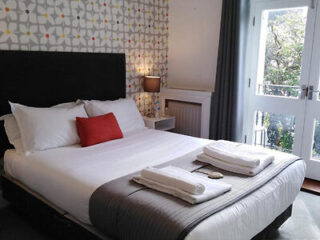 A neatly made bed with white sheets, gray throw, and red accent pillow. Two sets of folded towels are placed on top. The room features patterned wallpaper and a lamp beside a window with curtains.