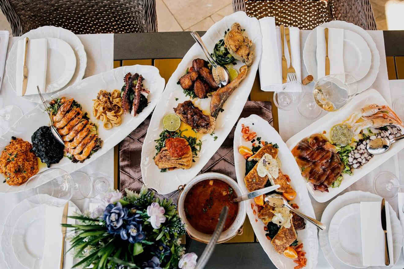 A table with various plated dishes, including grilled meats, seafood, and sides, set with utensils and napkins. A centerpiece with flowers is visible, along with empty wine glasses.