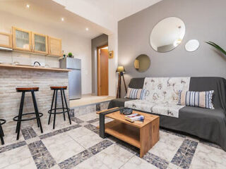 Modern living room with tiled flooring, a patterned sofa, and a wooden coffee table. Bar stools line a counter near the kitchen area. Round mirrors hang on the wall.