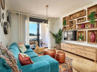 A cozy living room with a teal sofa, wooden shelves, potted plants, round ottomans, and a TV. Large windows lead to a balcony with outdoor seating. Bright and inviting decor.