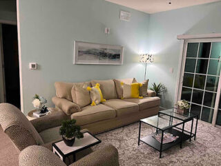 A living room with a beige sofa, yellow cushions, a patterned armchair, glass coffee table, potted plants, a standing lamp, wall art, and a sliding glass door.