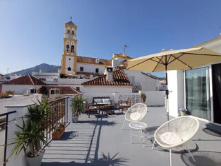 Rooftop terrace with chairs, tables, and a large umbrella. View of a church tower and mountain in the background. Clear blue sky.