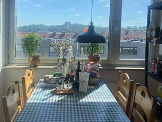 Dining table with a checkered tablecloth, surrounded by chairs, has various items including plants, condiments, and a pendant lamp above. Windows show a view of rooftops and sky.