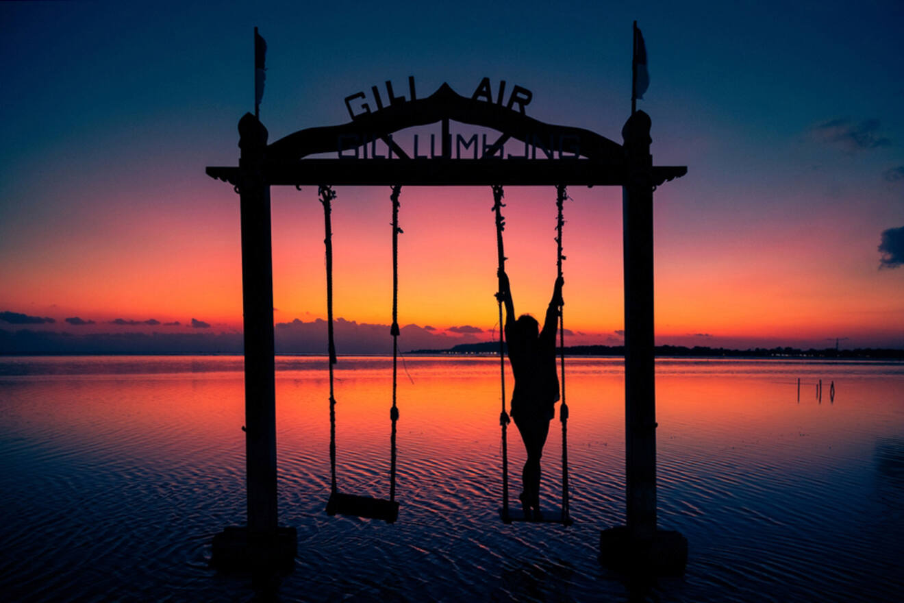 Silhouette of a person on a swing at sunset over water, with a sign overhead reading "Gili Air, Lombok, Indonesia.