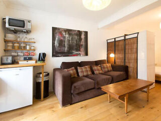 A living room with a brown sofa, plaid cushions, wooden coffee table, mini fridge, microwave, shelves with jars, artwork on the wall, and a room divider.